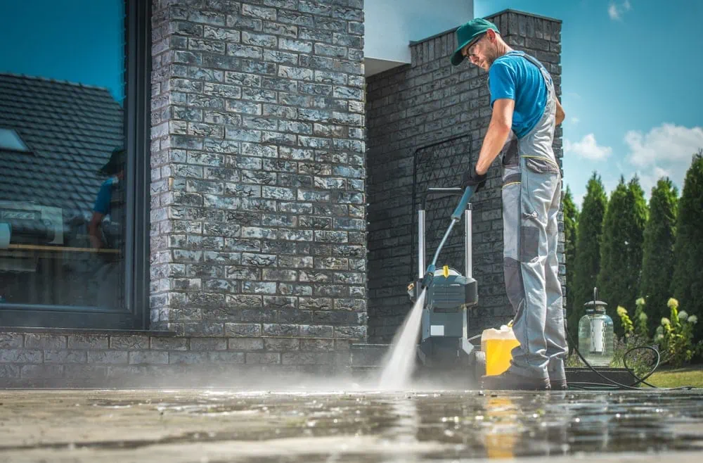 A worker pressure washing a paved surface beside a home